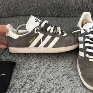 Grå Adidas Gazelle sneakers - Säljer ett par klassiska Adidas Gazelle sneakers i grå mocka med vita detaljer och vita ränder på sidorna. Skorna har snörning, platt sula och rund tå. Perfekta för en stilren och sportig look. Sitter ett par extra snören från saint Laurent eftersom jag tycker det blir otroligt mycket snyggare!🤟🏽