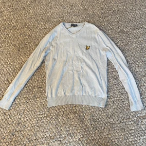 Ljusgrå v-ringad tröja från Lyle & Scott - Ljusgrå v-ringad tröja från Lyle & Scott med klassisk gul logga på bröstet. Tröjan har långa ärmar och ribbade muddar vid ärmslut och nederkant. Perfekt för en stilren och enkel look.