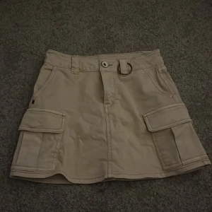 Beige cargokjol med fickor - Säljer en beige cargokjol med stora fickor på sidorna och bälteshällor i midjan. Kjolen har en O-ring detalj framtill och är i en medellång modell. Perfekt till sommaren😋Köpt från kids brand store