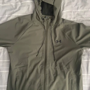 Olivgrön vindjacka från Under Armour - Säljer en olivgrön vindjacka från Under Armour med huva och dragkedja framtill. Jackan har två sidofickor och justerbar dragsko vid huvan. Perfekt för utomhusaktiviteter och träning.