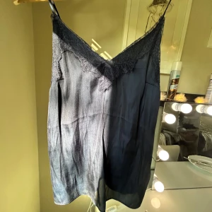 Mörkblå linne med spetsdetaljer - Säljer en mörkblå linne med tunna axelband och v-ringning. Linnet har fina spetsdetaljer längs urringningen och är tillverkad i ett lätt, glansigt material som ger en elegant känsla.