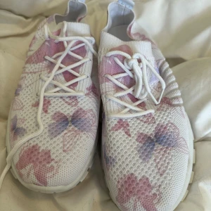 Vita sneakers med blommigt mönster - Säljer ett par vita sneakers med rosa och lila blommigt mönster. Skorna har snörning och är tillverkade i ett ventilerande syntetmaterial med meshstruktur. Perfekta för dig som vill ha en sportig och färgglad look. Storlek 40
