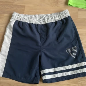 Marinblå badshorts med vita detaljer från East Side - Snygga marinblå badshorts med vita ränder och vit midja. Har elastisk midja och tryckt logga på ena benet. Perfekta för stranden eller poolen.