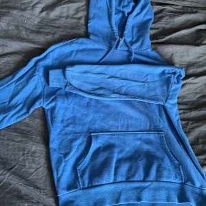 Blå hoodie från H&M - Säljer en klassisk blå hoodie från H&M med huva och magficka. Perfekt för en avslappnad stil. Tröjan har långa ärmar och är gjord i mjukt material som känns skönt mot huden.