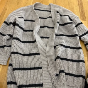 Beige och svart randig stickad kofta - Mysig stickad kofta i beige med svarta horisontella ränder. Koftan har lång ärm och öppen framtill utan knäppning. Perfekt för lager-på-lager och passar till många olika outfits.