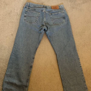 Blå jeansbyxor från Crocker (made in Italy) - Klassiska blå jeans från Crocker (tillverkade i Italien) med raka ben och normal passform. Byxorna har fem fickor, dragkedja och knapp framtill samt detaljerade nitar. Tillverkade i slitstarkt denimtyg och har en tidlös look som funkar till det mesta. (Har en nästintill osynlig fläck på framsidan av högerbenet)