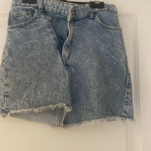 Blå jeanskjol från H&M - Säljer en blå jeanskjol från H&M med råa kanter och klassisk femficksdesign. Kjolen har bälteshällor och dragkedja framtill. Perfekt för en avslappnad stil.