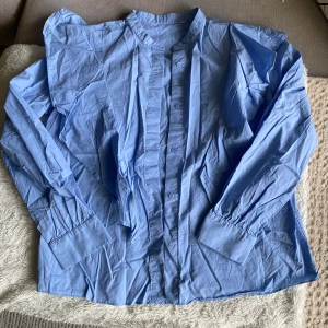 Ljusblå skjorta med volangdetaljer - Säljer en ljusblå skjorta med volangdetaljer längs axlarna och bröstet. Skjortan har lång ärm, knappar framtill och klassisk krage. Skjortan är från & Other Stories men lappen är tyvärr bortklippt. Superfint skick och sparsamt använd. I storlek 36. 