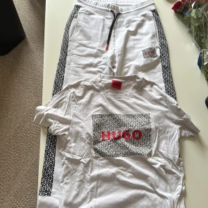 Hugo Boss med logga - Vit t-shirt från Hugo Boss med stor röd HUGO-logga på bröstet och svartvitt mönster runt loggan. Klassisk passform och rund halsringning. Perfekt för dig som gillar stilrena och trendiga plagg.