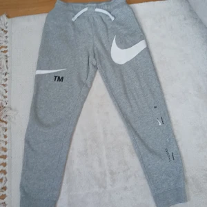 Grå mjukisbyxor från Nike - Säljer ett par grå mjukisbyxor från Nike med stora vita swoosh-loggor och tryckdetaljer på benen. Byxorna har dragsko i midjan och resår vid bensluten. Perfekta för en avslappnad stil.