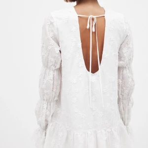 Vit blommig klänning med öppen rygg och volang - Superfin vit klänning med broderade blommor, volang nertill och långa puffärmar i transparent material. Öppen rygg med knytband upptill ger en extra detalj. Perfekt för sommarens alla tillfällen!