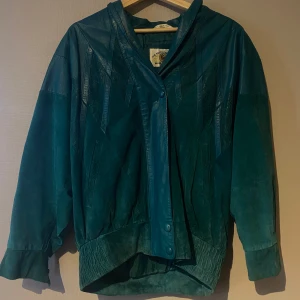 Turkos skinnjacka med knappar - Säljer en turkos skinnjacka med knäppning framtill och bred krage. Jackan har långa ärmar och en något oversized passform. Snygga detaljer i skinnet ger jackan en unik look. En mix av material: mocha och läder. 