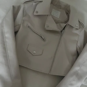 Beige croppad skinnjacka med dragkedja🤍 - Säljer en beige croppad skinnjacka med klassisk bikerkrage, silverfärgade dragkedjor och knappdetaljer. Jackan har lång ärm och en snedställd bröstficka med dragkedja samt en ficka med lock och knapp framtill. Säljer den pågrund av att det inte är min stil har aldrig använt den och den ligger på ungefär 120-200kr pris kan såklart diskuteras💕