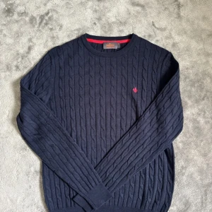 Mörkblå kabelstickad tröja från Morris - Säljer en mörkblå kabelstickad tröja från Morris med rund halsringning och diskret röd logga broderad på bröstet. Tröjan har långa ärmar och ribbade muddar vid ärmslut och nederkant. Perfekt för en stilren look. Passar perfekt med en skjorta under. Storlek M