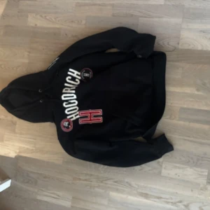 Svart hoodie från Hoodrich med tryck - Säljer en svart hoodie från Hoodrich med vit och röd text samt logga på framsidan. Tröjan har huva och en stor ficka framtill. Perfekt för dig som gillar streetwear och vill ha något bekvämt och snyggt.💞