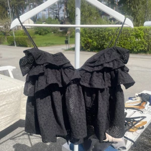 Svart volangtopp - Säljer en svart axelbandslös topp med volanger och broderade hål. Toppen har tunna axelband och är perfekt för sommardagar. Snygg och luftig modell med fina detaljer.