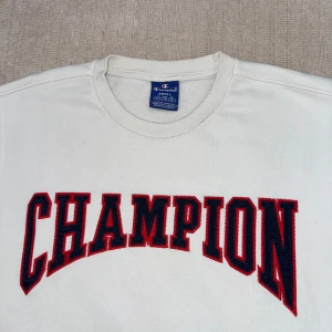 Vit sweatshirt från Champion - Vit sweatshirt från Champion med stort broderat CHAMPION-tryck i rött och mörkblå på bröstet. Klassisk rund halsringning och ribbade muddar. Liten Champion-logga på ärmen.