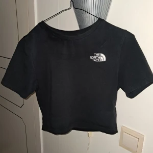 Svart croppad t-shirt från The North Face - Säljer en svart croppad t-shirt från The North Face med liten vit logga på bröstet. T-shirten har korta ärmar och är tillverkad i mjuk bomull. 