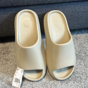 Beige Yeezy Slides från Adidas - Säljer ett par beige Yeezy Slides från Adidas. De har en minimalistisk design med öppen tå och platt sula, tillverkade i syntetmaterial. Perfekta för en avslappnad stil och riktigt bekväma att ha på sig. De är bara testade och inte använda 