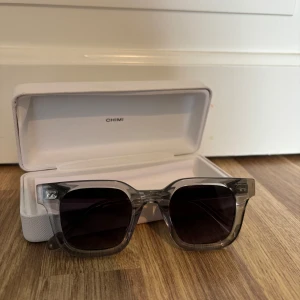 Grå transparenta solglasögon från CHIMI 04 - Snygga solglasögon från CHIMI med transparent grå båge och mörka fyrkantiga linser. Bågarna är i plast och har en modern, stilren design. Kommer med ett vitt CHIMI-fodral.
