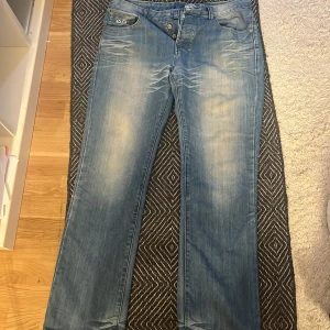 Blå jeansbyxor med broderade detaljer - Säljer ett par blå jeansbyxor från VB med snygga broderade detaljer på bakfickan och knappstängning fram. Byxorna har normal passform och klassisk femficksdesign. Perfekta för dig som gillar en avslappnad men stilren look. Inte använt dom en endå gång:))