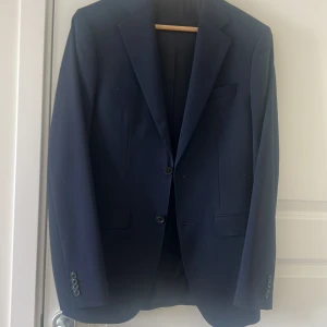 Mörkblå kostym med vit skjorta - Säljer en klassisk mörkblå kostym med matchande kavaj och byxor samt en vit skjorta. Kavajen har två knappar och fickor med lock. Perfekt för bal eller fest. Skjortan är långärmad med krage och knäppning framtill. Bara använd en gång och är ny kemtvättad.