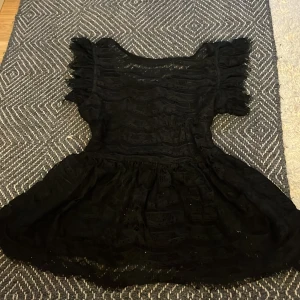 Svart spetströja från Gina Tricot - Supersöt svart tröja från Gina Tricot med volangdetaljer och spetsmönster. Klänningen har korta ärmar och en lätt utställd kjol. Perfekt för dig som vill ha en feminin och trendig look.