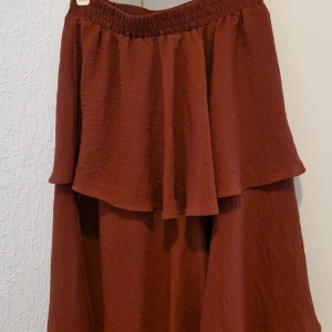 Rostbrun kjol med volang från Gina Tricot(oanvänd) - Säljer en rostbrun kjol från Gina Tricot med bred volang upptill och resår i midjan. Kjolen har en mjuk, lätt struktur och är perfekt för dig som gillar en flowy look. Har fått present, passade ej.