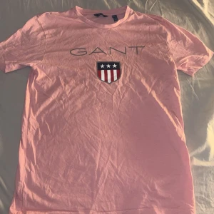 Rosa t-shirt från GANT med logga - Säljer en rosa t-shirt från GANT med tryckt logga på bröstet. korta ärmar. Perfekt för en avslappnad och stilren look. Använd en gång. Hör av er vid frågor.