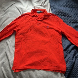 Röd pikétröja från Polo Ralph Lauren - Säljer en klassisk röd pikétröja från Polo Ralph Lauren med blå broderad logga på bröstet. Tröjan har krage, knappar vid halsen och långa ärmar. Perfekt för en stilren och sportig look.