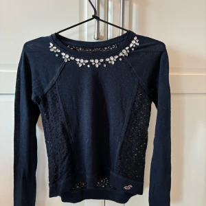 Mörkblå topp med spets och stenar från Hollister - Supersnygg mörkblå topp från Hollister med långa ärmar, spetsdetaljer på sidor och rygg samt glittriga stenar vid halsringningen. Perfekt för dig som vill sticka ut lite extra!