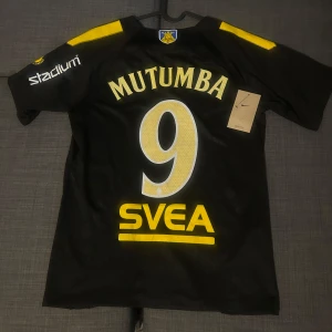 AIK fotbollströja Mutumba 9 - Säljer en svart AIK fotbollströja med gula detaljer, spelarnamn Mutumba och nummer 9 på ryggen. Tröjan har korta ärmar, AIK:s klubbmärke på bröstet och sponsorer i gult. Materialet är lätt och andas, perfekt för match eller träning. Passar som M men även S.