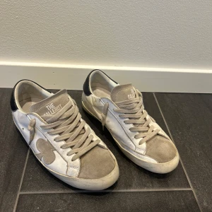 Vita och beige sneakers från The Gallerist - Snygga sneakers i vitt och grått med svarta detaljer på hälen och en dekorativ stjärna på sidan. Skorna har snörning och rund tå, samt en platt sula. Perfekta för en avslappnad och trendig stil. Skorna kommer utan original box.