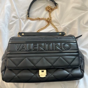Svart quiltad axelväska från Valentino - Säljer en svart quiltad axelväska från Valentino med guldig kedja och tydlig logga framtill. Väskan har ett lock med lås och snygga detaljer. Perfekt för dig som vill ha en stilren och trendig accessoar.