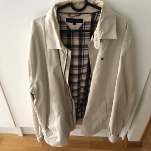 Beige jacka från Tommy Hilfiger - Snygg beige jacka från Tommy Hilfiger med klassisk krage och dragkedja. Insidan har rutigt foder i beige, rött och blått. Liten broderad logga på bröstet. Perfekt för övergångssäsonger.