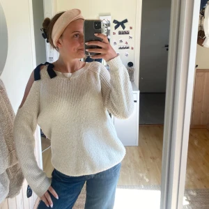 Vit stickad tröja med rosett-detaljer - Säljer en vit stickad tröja med öppna axlar och mörkblå rosetter på axlarna. Tröjan har lång ärm och en avslappnad passform, perfekt för mysiga dagar.