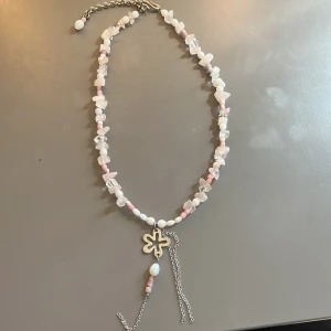 Halsband med pärlor och stenar i rosa och vitt - Handgjort halsband med oregelbundna pärlor och stenar i rosa och vitt, dekorerat med en silverfärgad blomformad detalj och kedjor. Justerbar längd med metallkedja och karbinhake. 