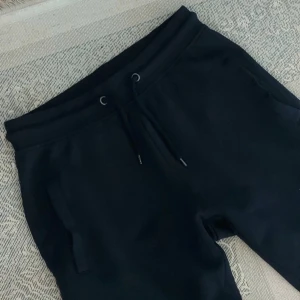 Svarta mjukisbyxor med dragsko - Svarta mjukisbyxor med elastisk midja och dragsko. Byxorna har två sidofickor och är tillverkade i mjukt material som passar perfekt för en avslappnad stil. Raka ben med mudd vid benslut.