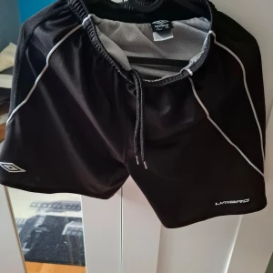 Svarta sportshorts från Umbro - Svarta sportshorts från Umbro med vita detaljer och snörning i midjan. Tillverkade i ett lätt och ventilerande polyestermaterial, perfekta för träning eller fotboll. Klassisk design med elastisk midja och logga på benet.