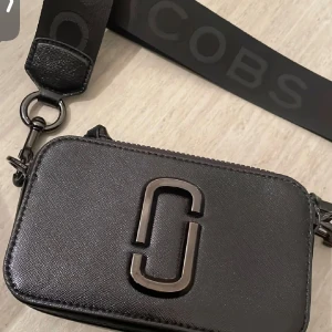 Svart axelväska från Marc Jacobs - Säljer en svart axelväska från Marc Jacobs med bred logotyp-prydd axelrem och dubbla dragkedjor. Väskan har en stilren design med metallogo framtill och är tillverkad i skinn. Perfekt för att bära det viktigaste med stil. Köptes för 4,500kr i NK. Knappt använd!!