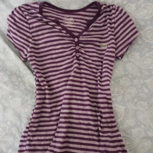 Lila y2k-topp från Lacoste - Assnygg topp från Lacoste med v-ringning och knappar framtill. Perfekt för y2k/downtownstil, älskar men måste tyvärr sälja då den är för liten 💝 öppen för prisförslag