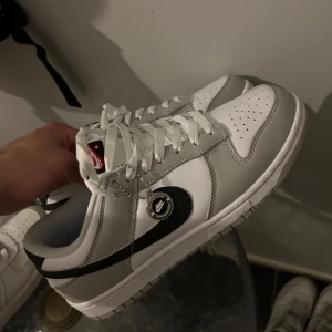 Nike Dunk Low i vitt och grått - Nike Dunk Low sneakers i vitt och ljusgrått med svart swoosh. Skorna har klassisk snörning, rund tå och platt sula. Detalj med Nike-tag på plösen och en liten kedja med Nike-logga på sidan. Perfekta för en stilren vardagslook.