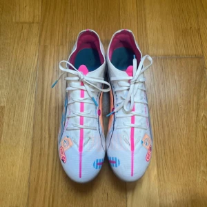 Vita puma fotbollsskor med färgglada detaljer - Säljer ett par vita fotbollsskor med snörning och färgglada detaljer i blått, rosa och orange. Skorna har rund tå, platt sula med dobbar och coola grafiska mönster på sidorna. Perfekta för dig som vill sticka ut på planen!