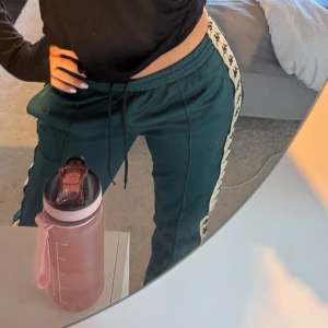 Gröna byxor från Kappa - Mörkgröna byxor från Kappa med vita sidor och svarta Kappa-loggor längs benen. Byxorna har resår och snörning i midjan samt raka ben. Perfekta för en sportig look.