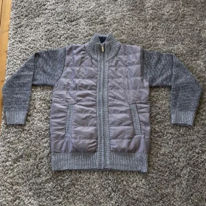 Grå stickad cardigan med dragkedja - Säljer en grå cardigan med stickade ärmar. Jackan har quiltad framsida i syntetmaterial och ribbade muddar vid ärmslut, krage och nederkant. Stängs med dragkedja framtill. Säljer eftersom den är för stor för mig, finns det frågor är det bara att ställa!