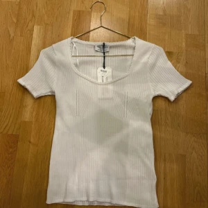 Vit ribbad topp från Holly & Whyte - Säljer en vit ribbad topp från Holly & Whyte med rundad halsringning och korta ärmar. Toppen är i mjukt material och har en figurnära passform. Perfekt till sommaren och enkel att matcha med olika outfits.