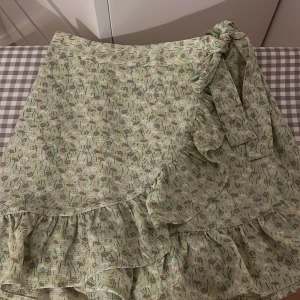 Grön blommig omlottkjol med volang -  ljusgrön omlottkjol med blommigt mönster och volangkant. Kjolen knyts i sidan med en rosett och har en luftig, somrig känsla. 