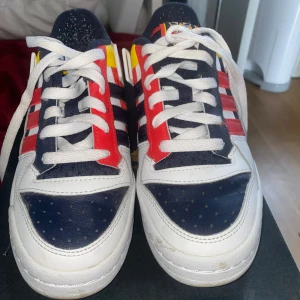 Adidas sneakers i rött, vitt och blått - Färgstarka Adidas sneakers med klassiska tre ränder i rött, vitt och marinblått. Skorna har detaljer i gult och svart, vit sula och snörning. Ovandelen är i skinn och textil, med rund tå och platt sula. Perfekta för dig som gillar retrostil och vill sticka ut.