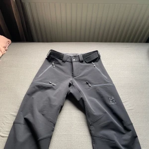 Svarta täckbyxor från Haglöfs - Säljer ett par svarta täckbyxor från Haglöfs med flera dragkedjeförsedda fickor och justerbar midja med kardborre. Byxorna har tryckknappar vid benslut och är gjorda i ett slitstarkt syntetmaterial, perfekta för utomhusaktiviteter och vinteräventyr.