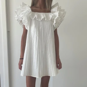 Vit plisserad kortklänning med volang - Superfin vit kortklänning med plisserat tyg och stora volanger runt axlar och halsringning. Använd 2 gånger! I super fint skick!!